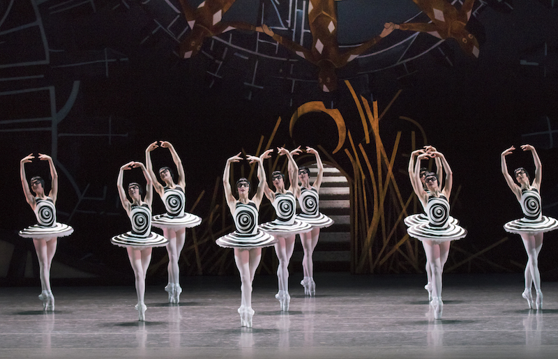 Nine ballerinas in black and white tutus that have a back swirling op art line on the front. All of the dancers don black page boy wigs and black glasses over their eyes